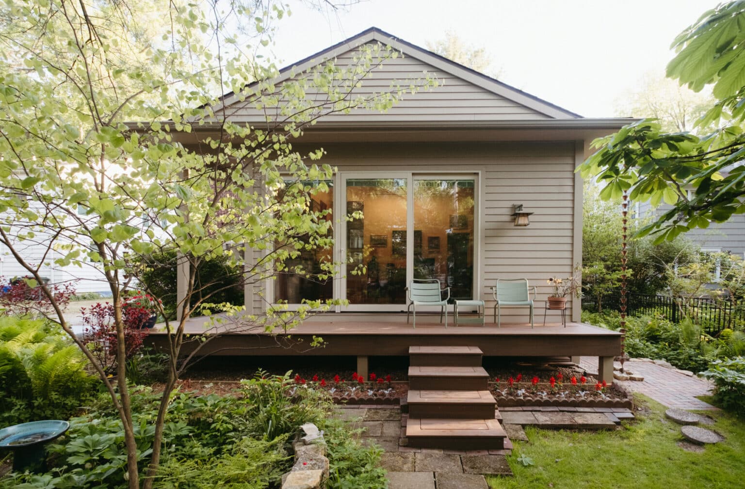 A small house with beige siding features a wooden deck, two chairs, a glass door, and steps leading to a garden with lush greenery and red flowers—an inviting Urbana IL home addition design.