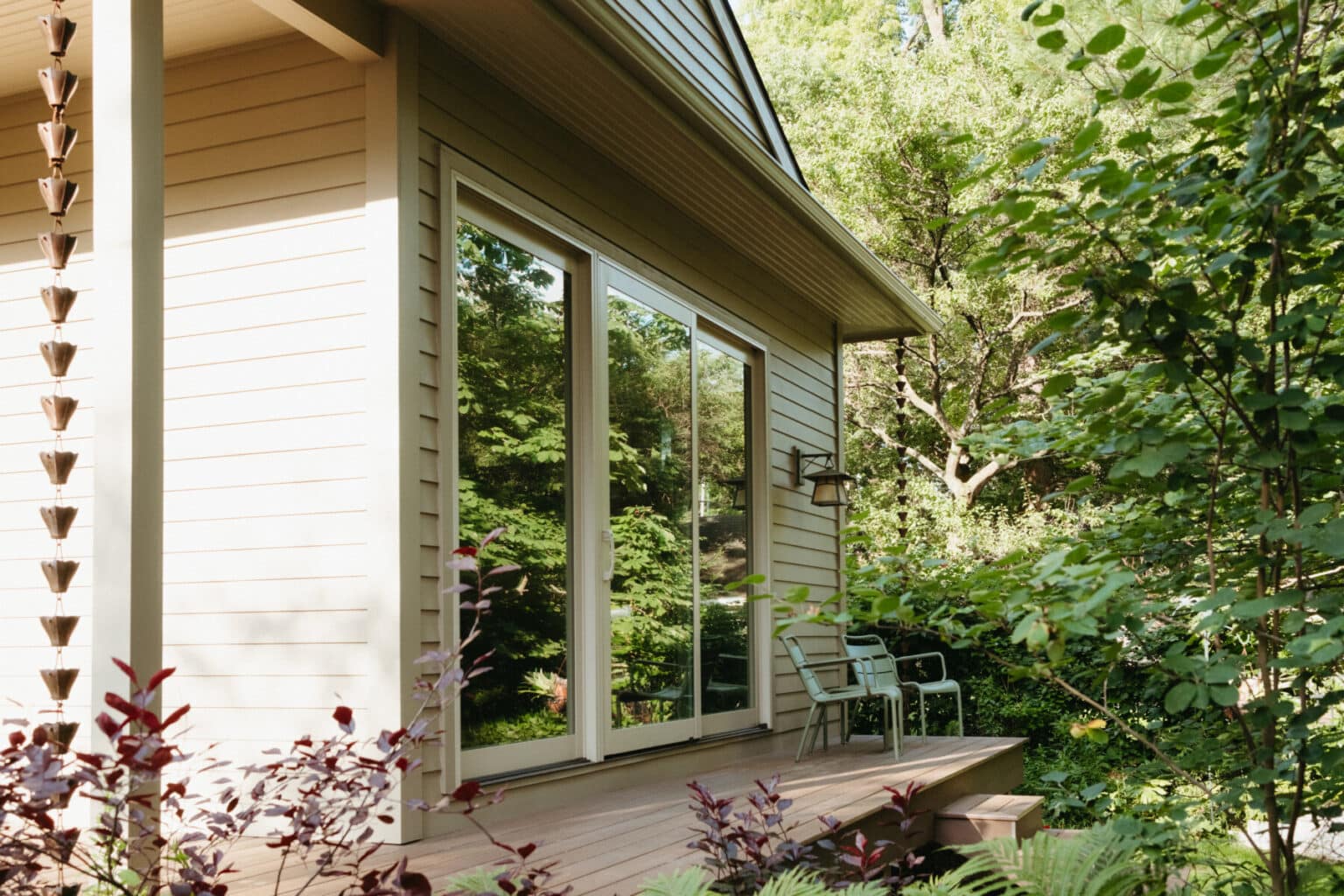 A beige house with sliding glass doors opens onto a small wooden deck with two chairs, surrounded by green trees and plants—showcasing thoughtful Urbana IL home addition design.
