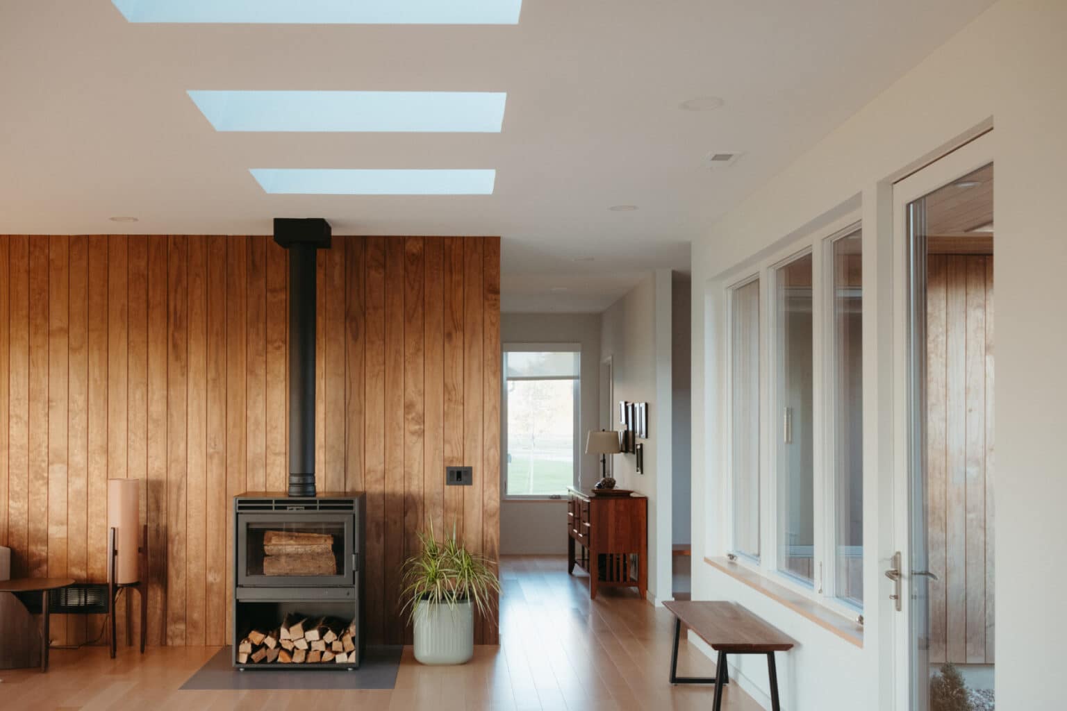 A modern living room reflects sleek modern house design Urbana IL, featuring wood-paneled walls, a black wood-burning stove, skylights, large windows, and minimal furniture including a bench and a small plant.