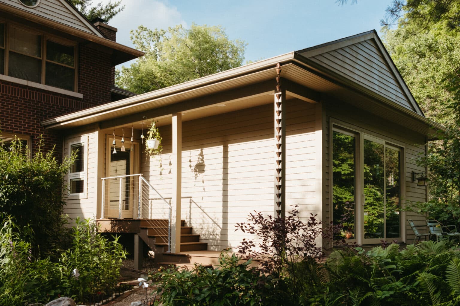A beige house extension with large windows and a small porch, surrounded by greenery and plants, showcases Urbana IL home addition design, seamlessly attached to a brick building.