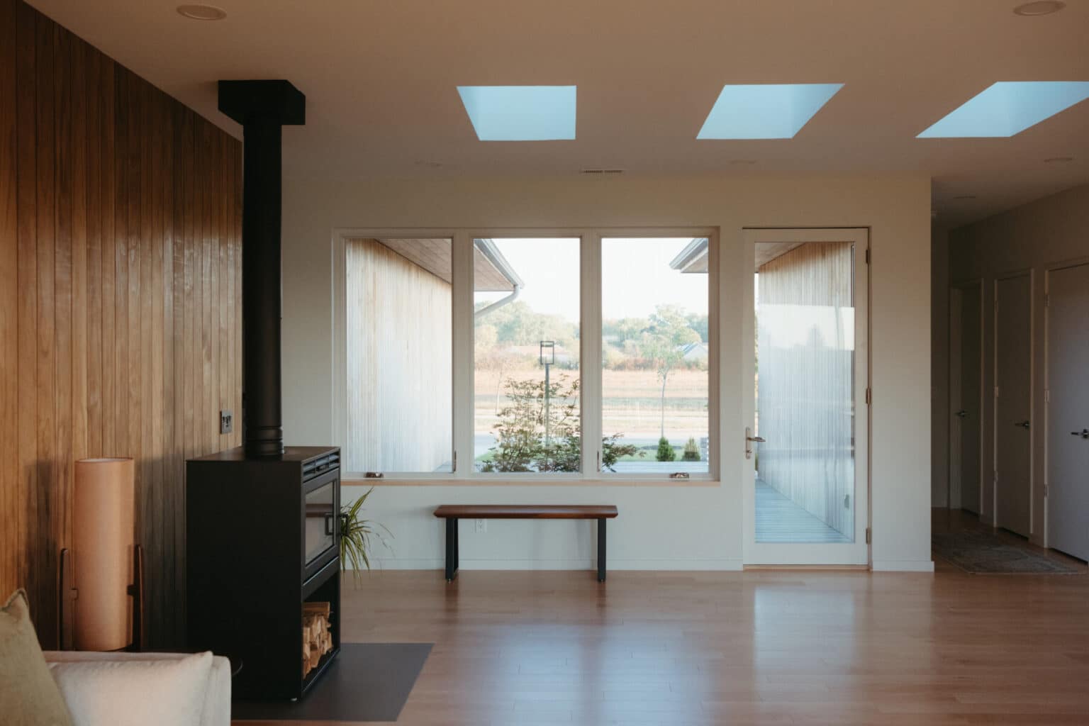 Minimalist living room in a modern house design Urbana IL, featuring wooden floors, a wood-burning stove, a bench under a large window, and skylights that fill the space with natural light. Glass door opens to the outdoors.