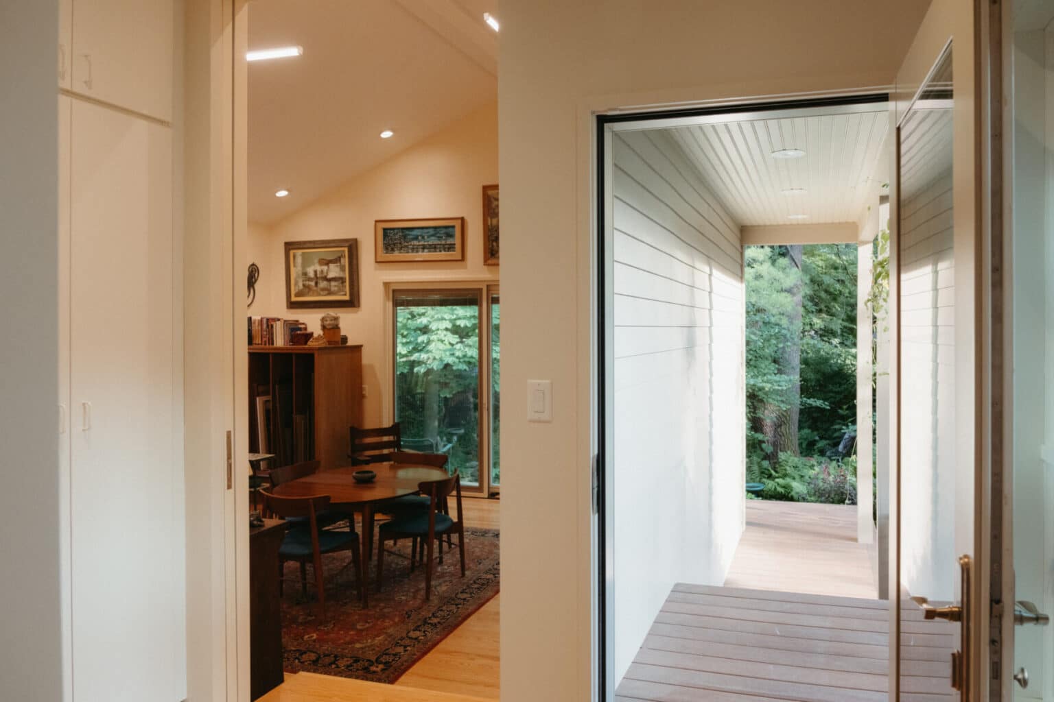 View from inside an Urbana IL home addition design, showing a hallway leading to a covered porch and a dining area with wooden furniture and bookshelves. Trees and greenery are visible outside.