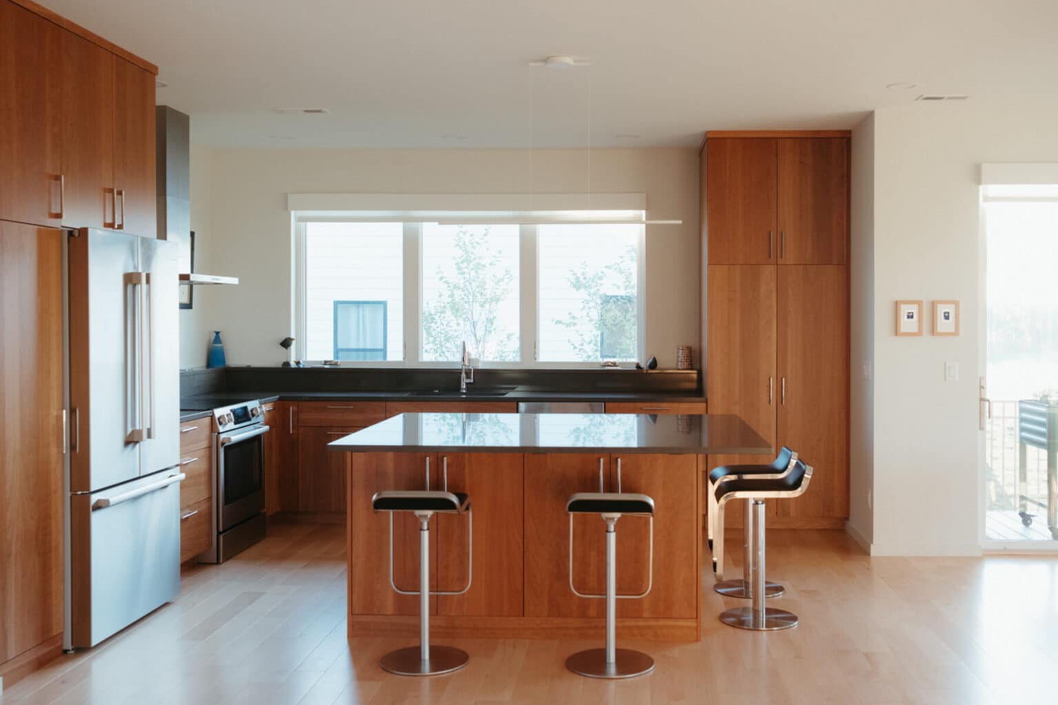 Modern kitchen with wooden cabinets, stainless steel appliances, a central island with black countertop, and three bar stools; large windows provide natural light—perfect for a modern house design in Urbana IL.