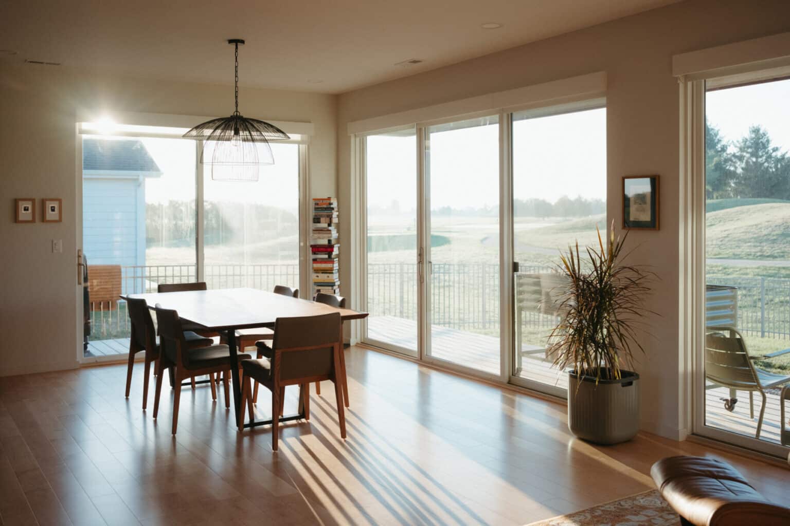 A dining area in a modern house design Urbana IL features a wooden table with six chairs, sunlit through large glass doors overlooking lush greenery. Nearby, a potted plant and bookshelf add charm to the inviting space.