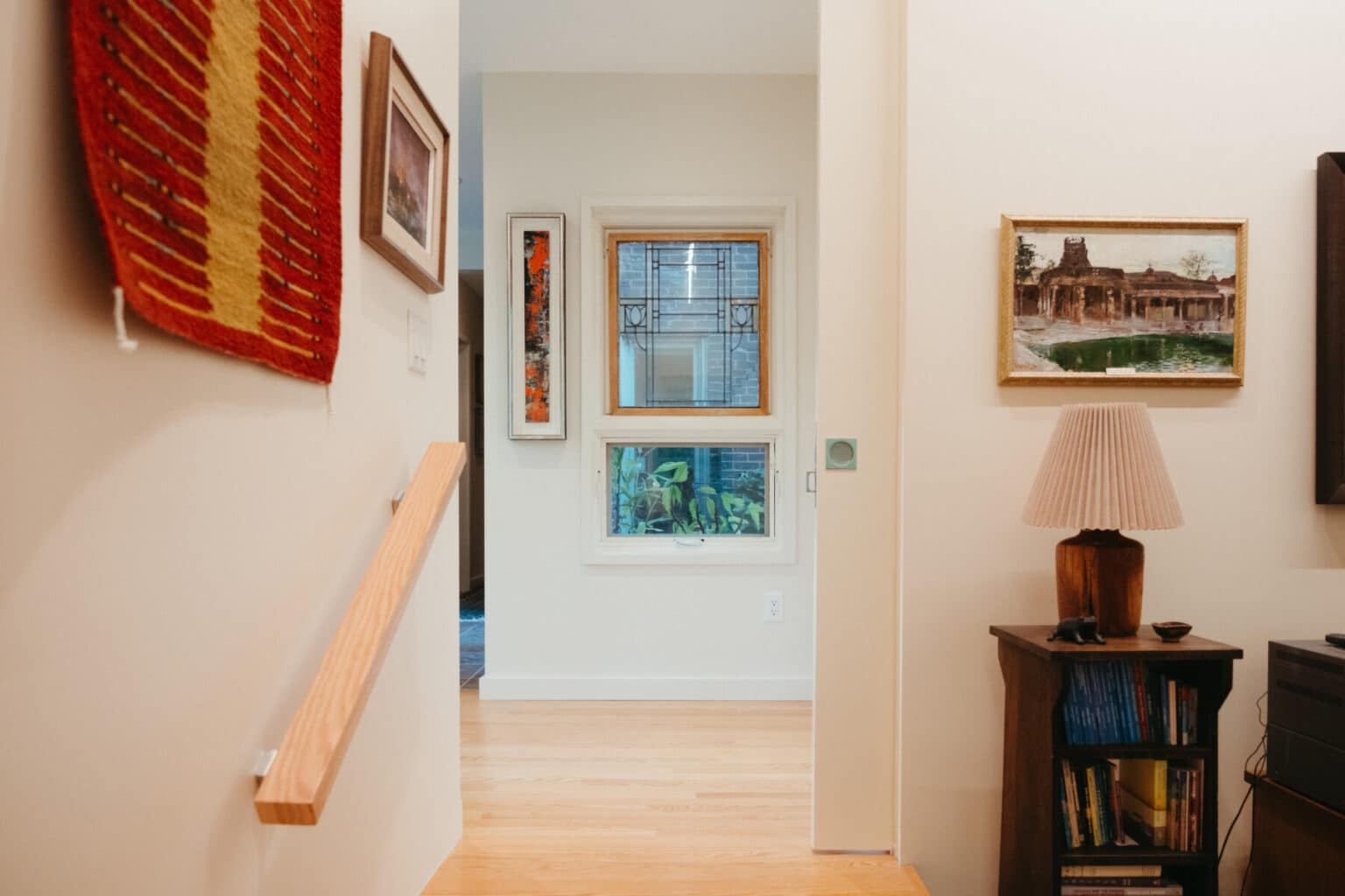 A well-lit hallway with wooden floors, decorated with framed art, a stained glass window, and a side table with a lamp and books—perfect inspiration for Urbana IL home addition design.