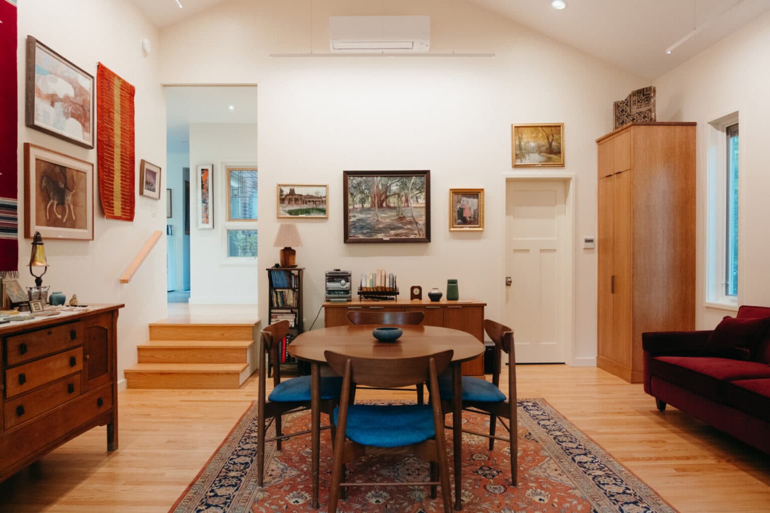 A dining area with a wooden table and four blue chairs, surrounded by various paintings, a rug, sideboard, bookcase, and red sofa in a well-lit room showcases stylish Urbana IL home addition design.