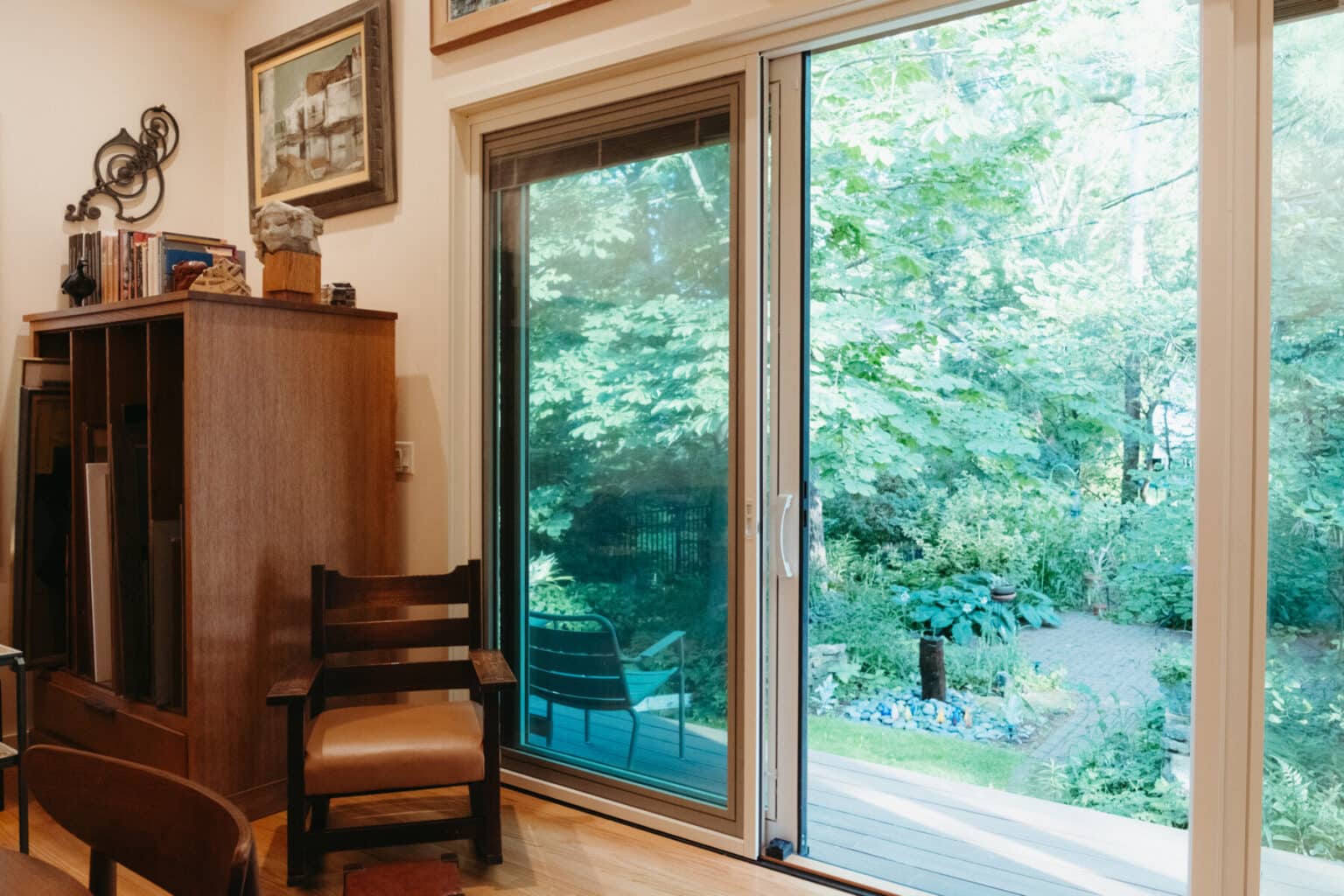 A wooden chair sits by a sliding glass door, part of an Urbana IL home addition design, opening onto a lush backyard garden with trees and a stone path.