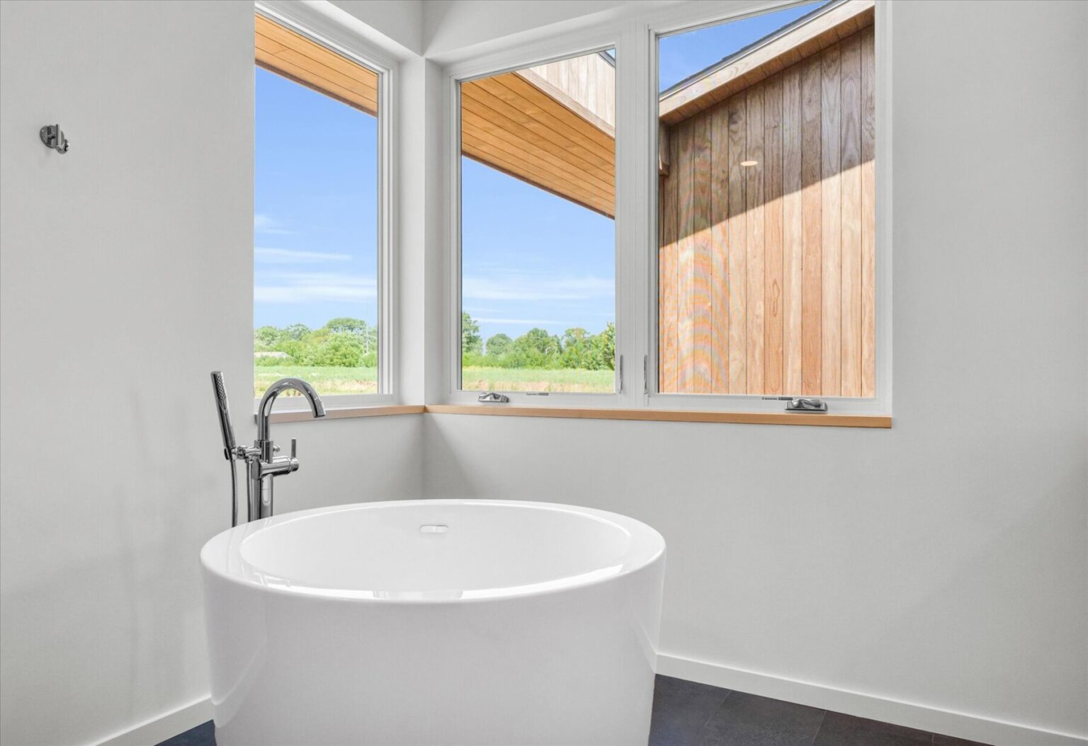 A modern white freestanding bathtub is positioned by large corner windows with views of greenery and a wooden exterior wall, reflecting the elegance of modern house design in Urbana IL.