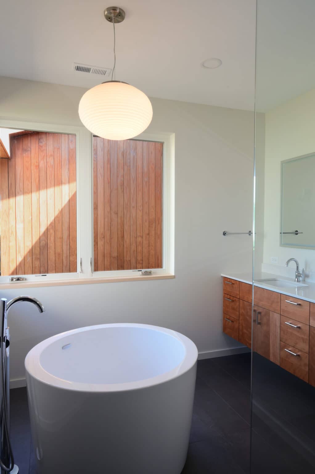 Modern bathroom with a freestanding white bathtub, wooden vanity with sink and mirror, pendant light, and two large windows framed by vertical wooden panels—perfect for a modern house design in Urbana IL.