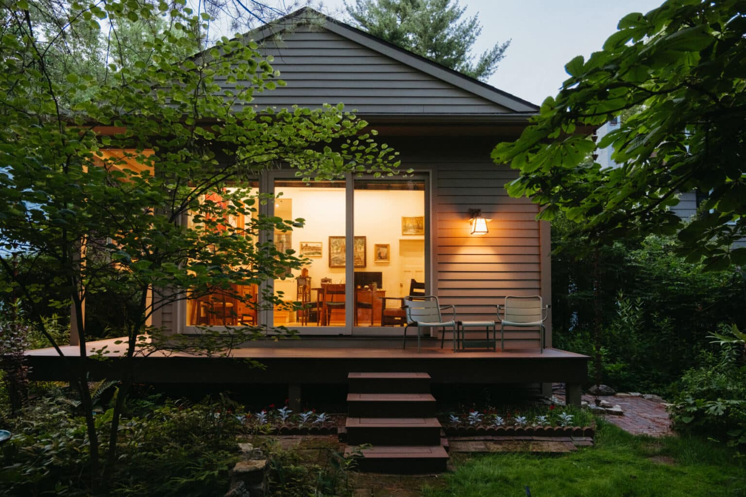 A small house with a lit interior is viewed from a garden. The porch has two chairs, and trees and plants surround the area, showcasing thoughtful Urbana IL home addition design.