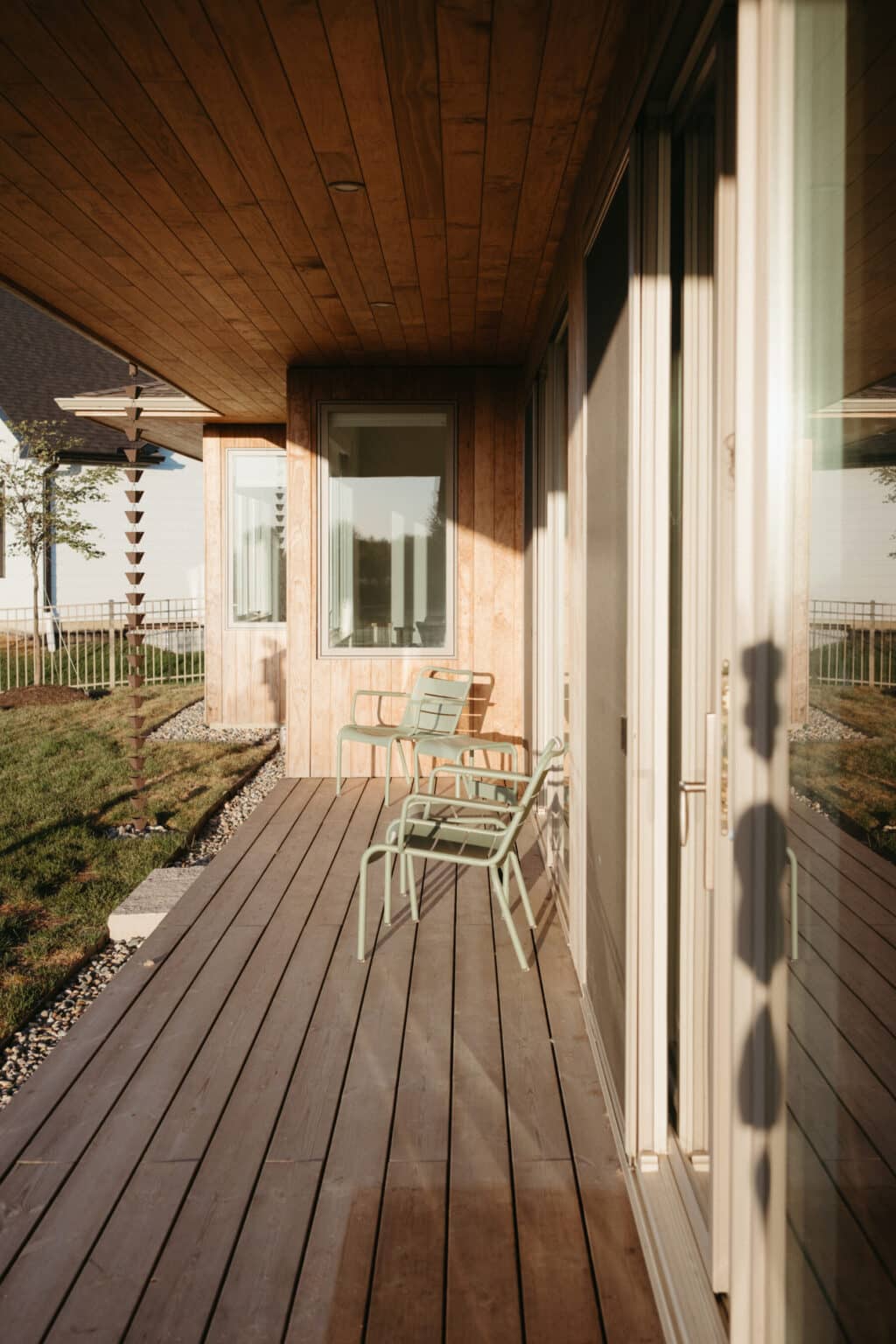 Two green metal chairs sit on a wooden deck outside a modern house design in Urbana, IL, with large windows and a wooden overhang, basking in the sunlight.