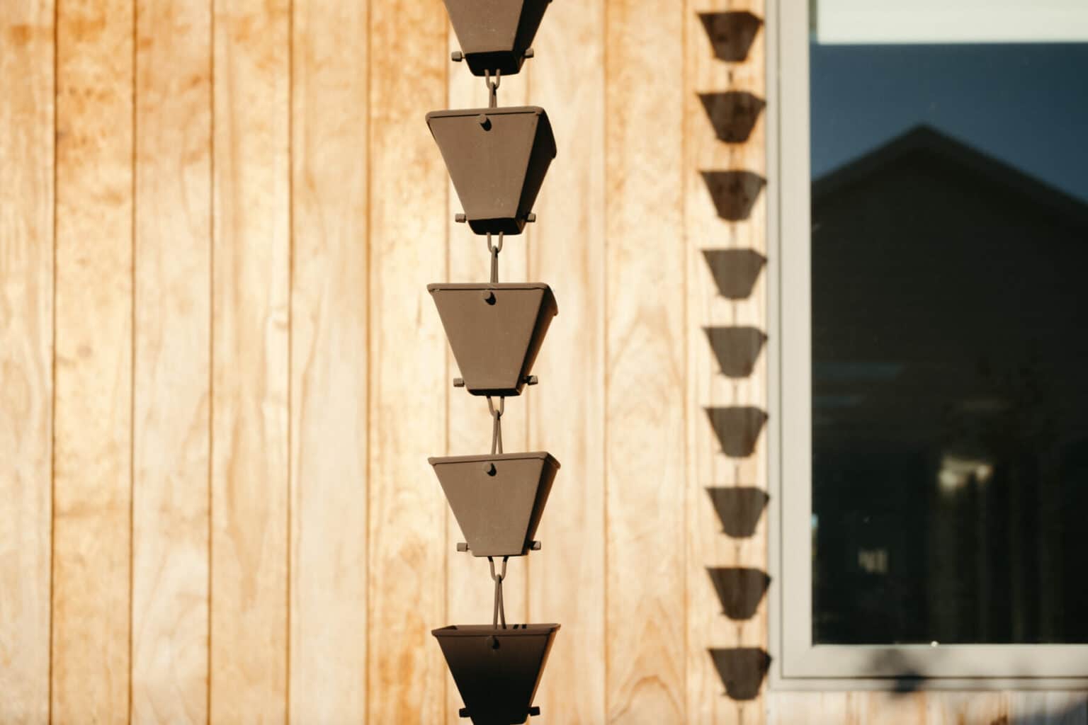 A vertical row of cone-shaped rain chain cups hangs in front of a wooden wall with a window to the right, casting distinct shadows—a striking accent for modern house design in Urbana IL.