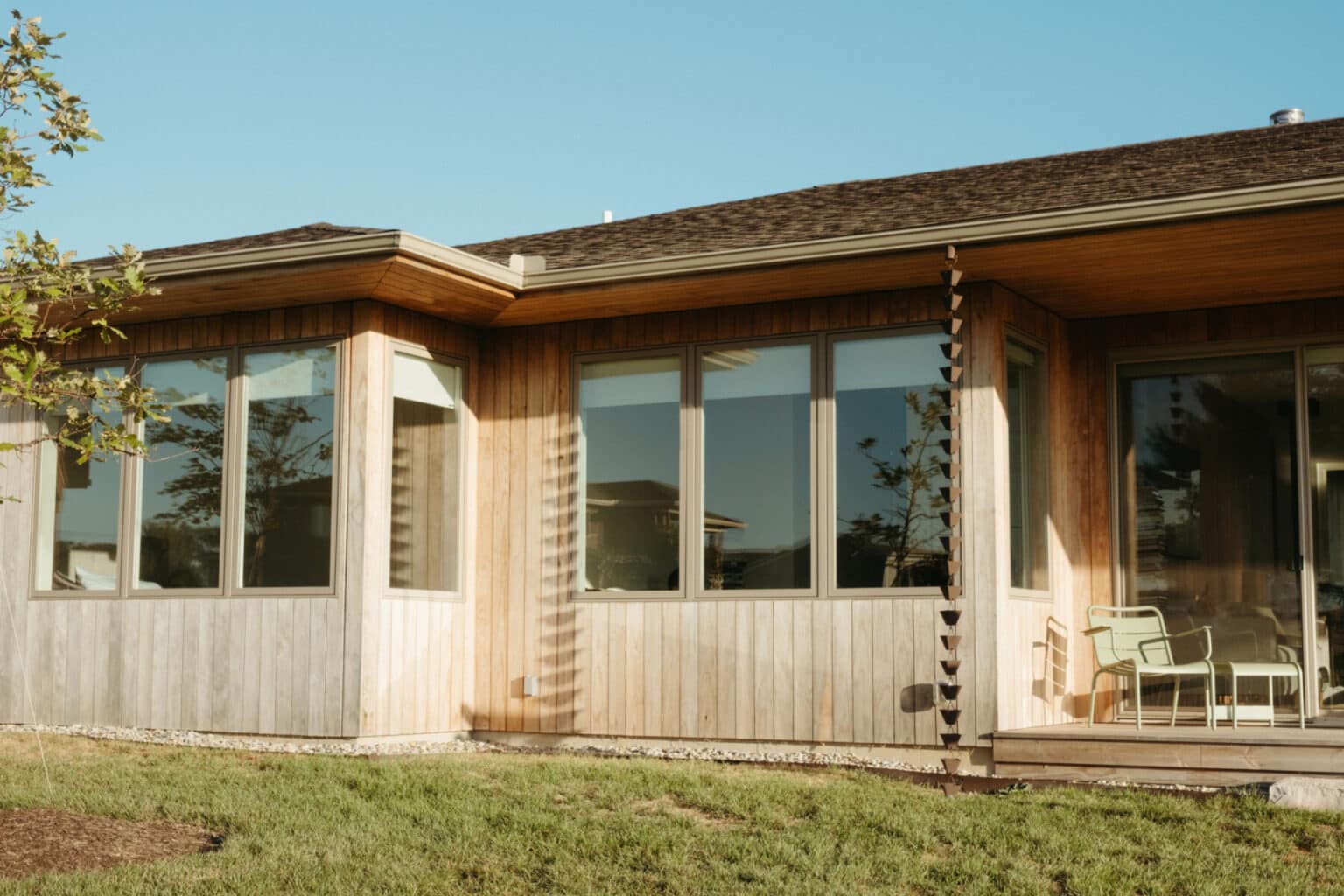 Single-story modern house design in Urbana, IL with large windows, wood siding, a rain chain, green lawn, and outdoor chairs on a small patio under a clear blue sky.