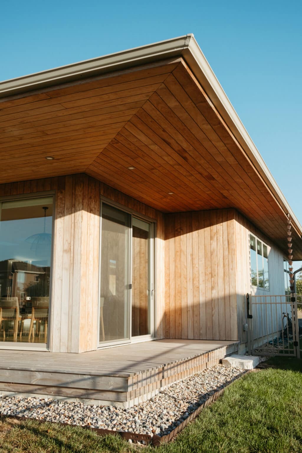 Modern wooden house exterior inspired by modern house design Urbana IL, featuring large sliding glass doors, a small porch with pebbles, and a well-maintained green lawn under a clear sky.