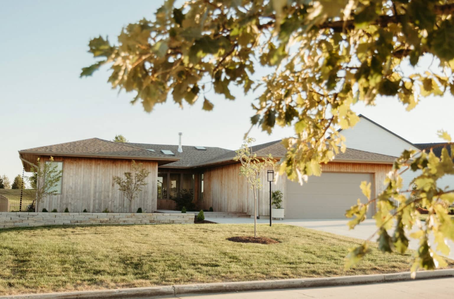 Single-story modern house design in Urbana IL with light wood siding, a two-car garage, landscaped front yard, and a tree in the foreground on a sunny day.