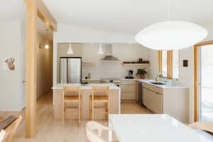 Modern kitchen with light wood floors, beige cabinets, an island with two chairs, stainless steel appliances, white pendant lights, and large windows—perfect for a mid century modern home renovation in Urbana IL.
