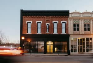 A two-story brick building with large front windows, offering downtown Champaign loft apartments and commercial space, stands next to a cream-colored building on a quiet street at dusk; blurred car lights streak by in front.