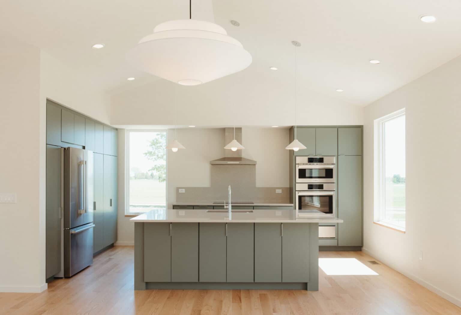 Modern kitchen with sage green cabinets, stainless steel appliances, a central island with sink, pendant lights, and large windows providing natural light—perfect for those seeking custom home design in Urbana IL.