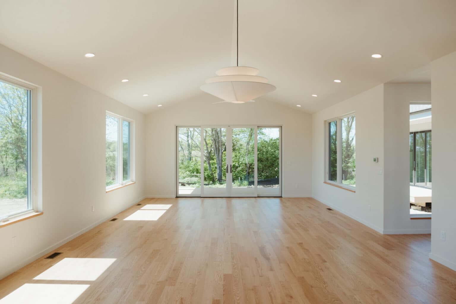 Bright empty room with light wood flooring, large windows, glass doors leading outside, recessed lighting, and a modern hanging light fixture—perfect for showcasing custom home design in Urbana IL.