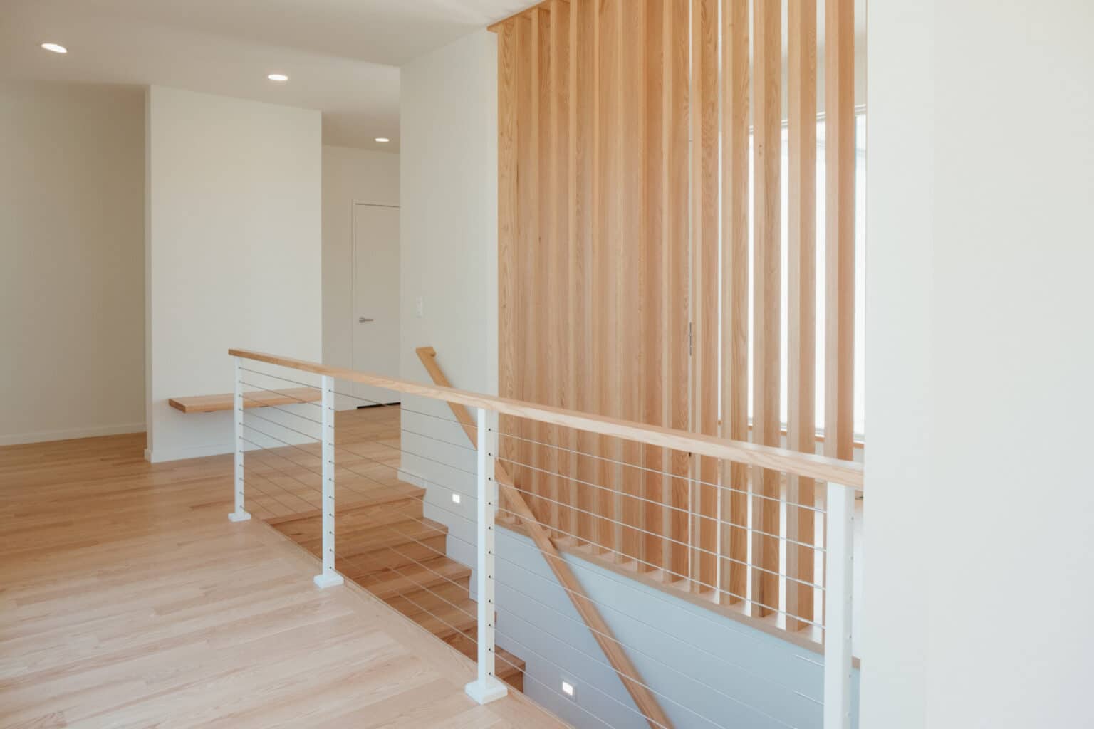 Modern interior with light wood floors, a staircase with cable railing, and a vertical slatted wood accent wall—showcasing the open, minimalist approach of custom home design in Urbana IL.