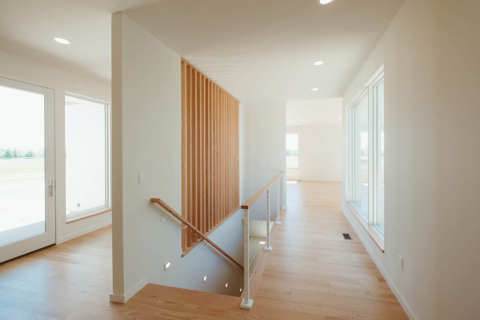 A modern hallway with light wood floors, white walls, large windows, and a staircase with wooden slats and a metal railing—showcasing the elegance of custom home design in Urbana IL.