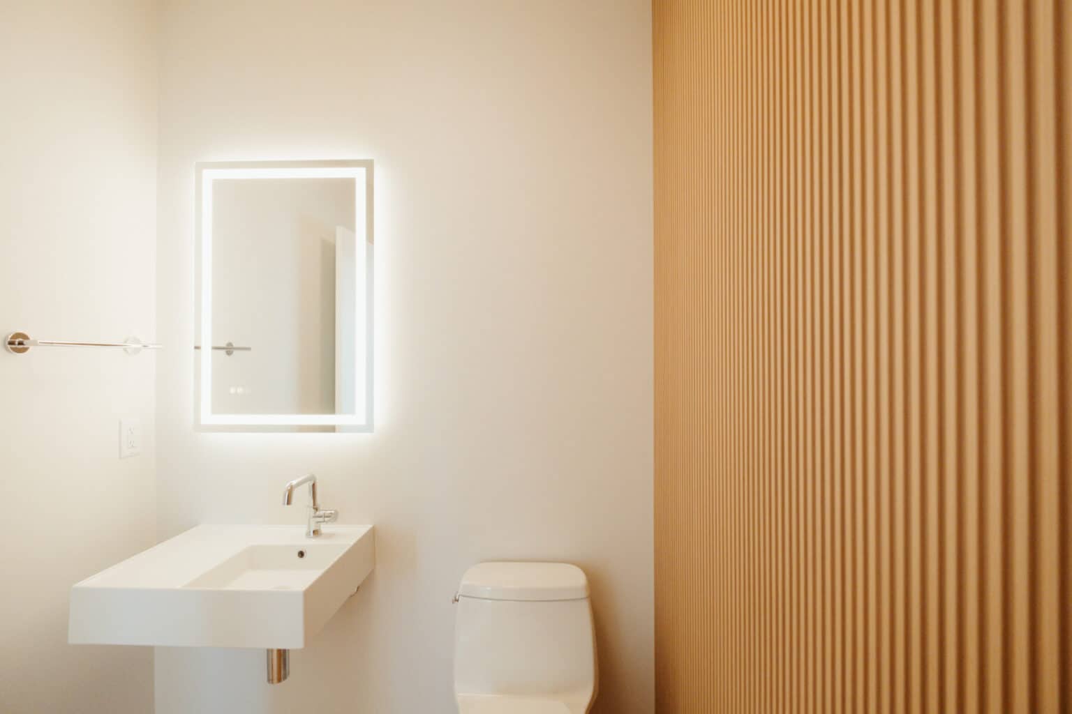 Minimalist bathroom with a rectangular sink, wall-mounted faucet, illuminated mirror, white toilet, and vertical wooden slat wall on the right—perfect for custom home design in Urbana IL.