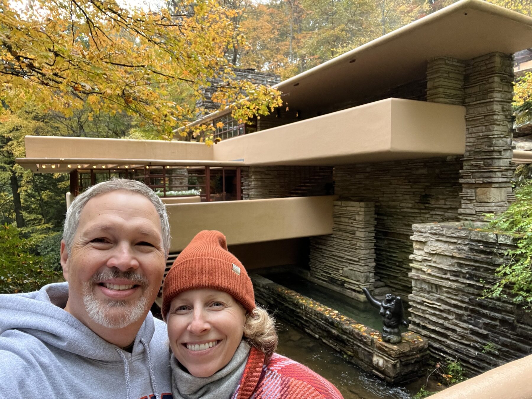 A smiling couple poses in front of Fallingwater, a modern house with stone and concrete terraces over a stream, surrounded by autumn trees.