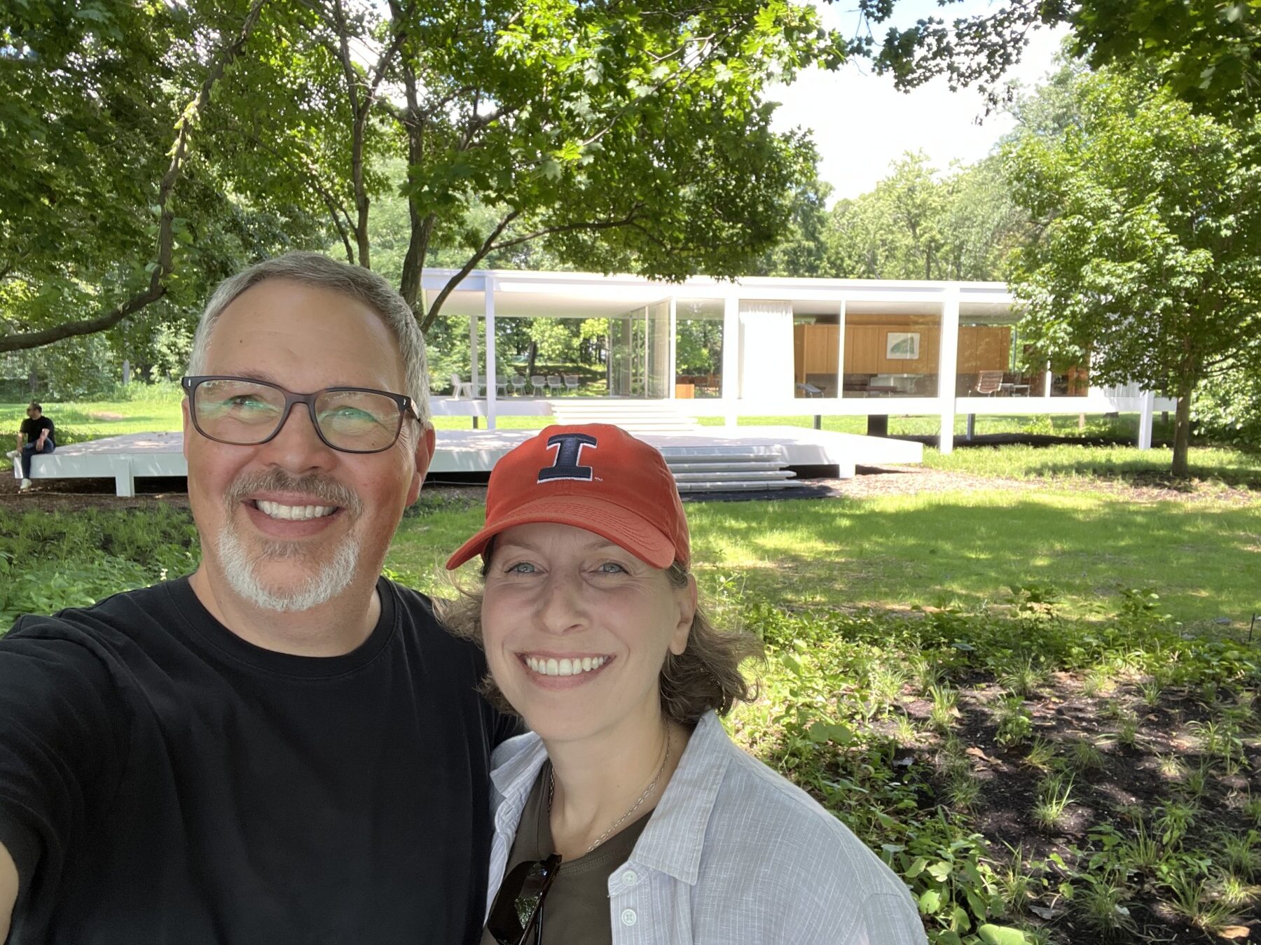 Two people smiling in front of the Farnsworth House, a modern glass house surrounded by trees and greenery on a sunny day, showcasing inspiration for any design build firm in Urbana IL.