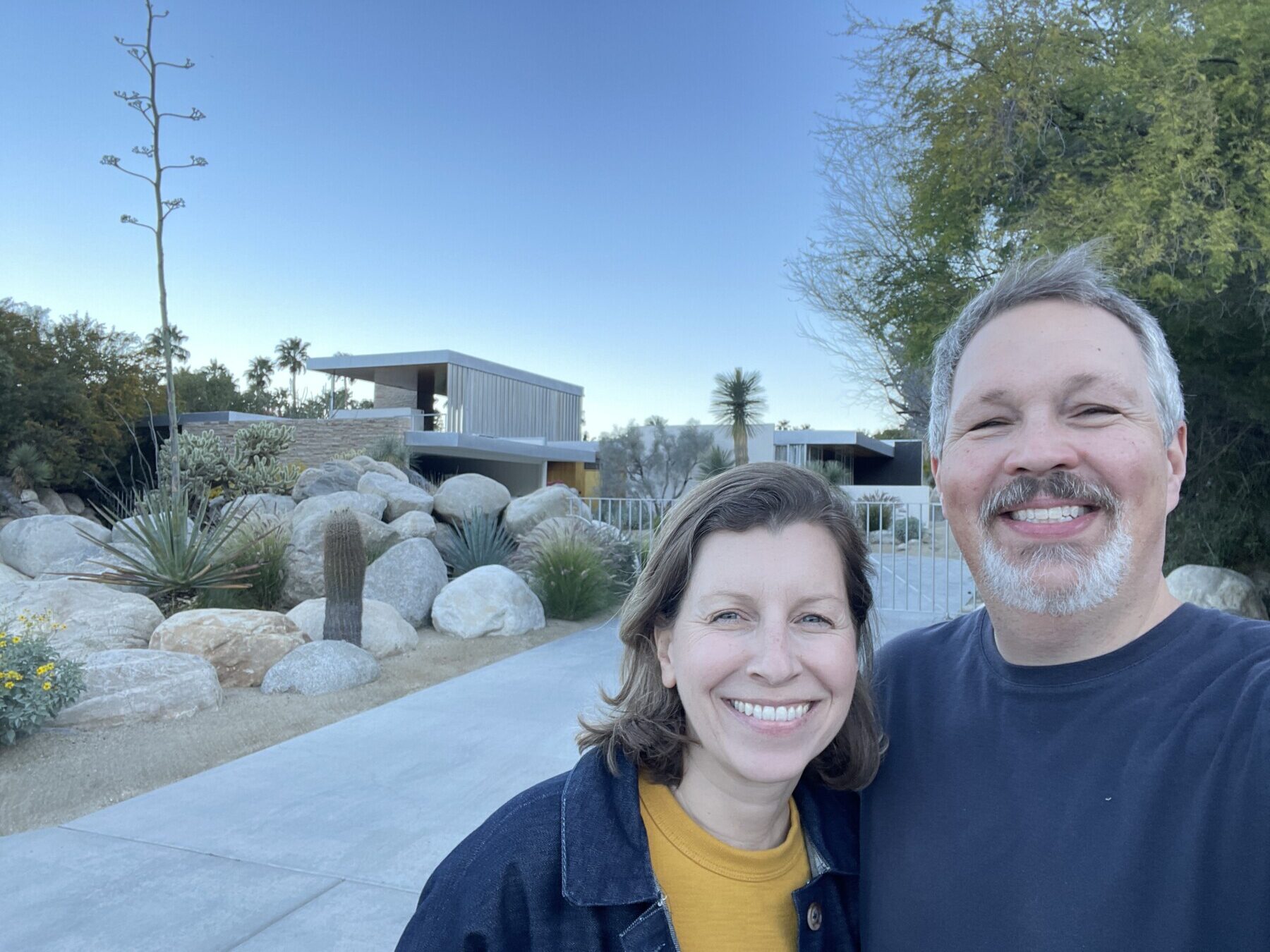 A smiling man and woman stand outside in front of a modern house with large rocks, desert plants, and clear blue sky.