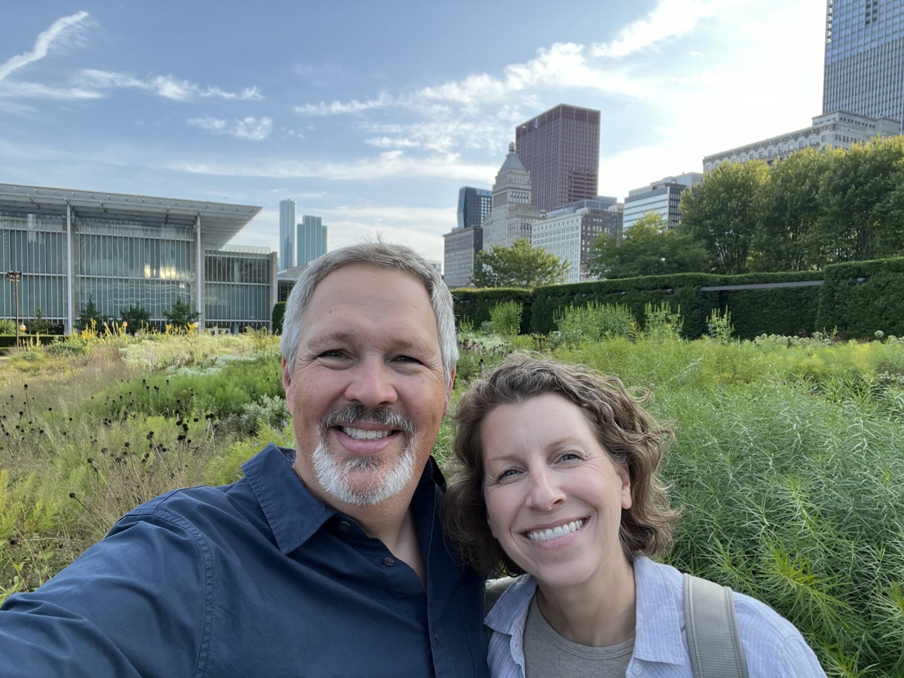 A man and woman smile for a selfie in a city park with greenery and tall buildings in the background under a partly cloudy sky.