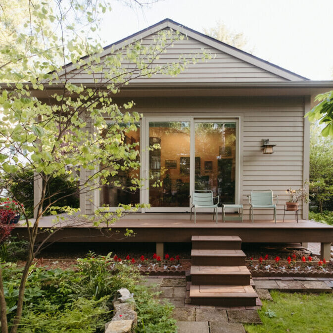 A small house with beige siding features a wooden deck, two chairs, a glass door, and steps leading to a garden with lush greenery and red flowers—an inviting Urbana IL home addition design.