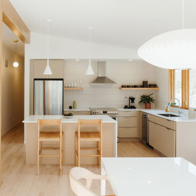 Modern kitchen with light wood floors, beige cabinets, an island with two chairs, stainless steel appliances, white pendant lights, and large windows—perfect for a mid century modern home renovation in Urbana IL.