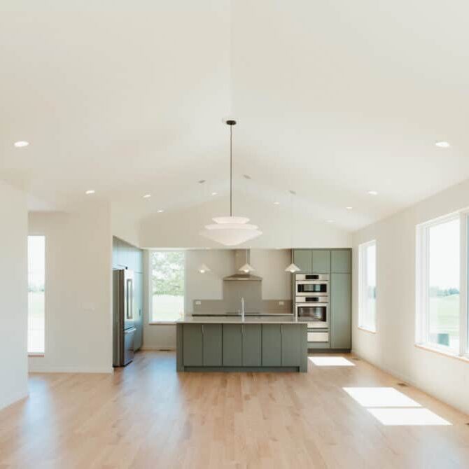 A modern, open-concept kitchen with light wood floors, sage green cabinets, stainless steel appliances, and large windows letting in natural light—perfect for a custom home design in Urbana IL.
